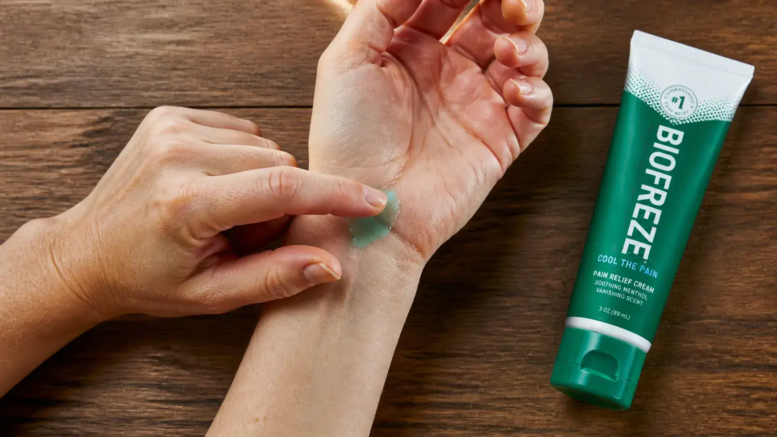 Hand applying cooling pain relief gel to the inside of a wrist, with the Biofreeze tube visible nearby on a wooden surface